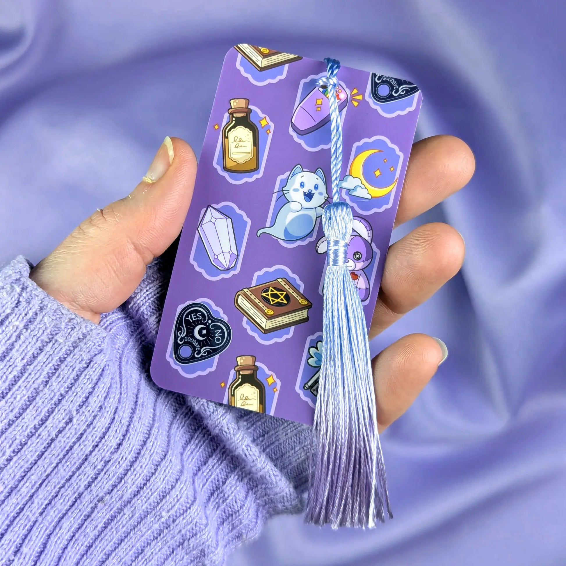 Purple bookmark with cute ghost hunter illustrations with tassel held by a hand against a matching purple background