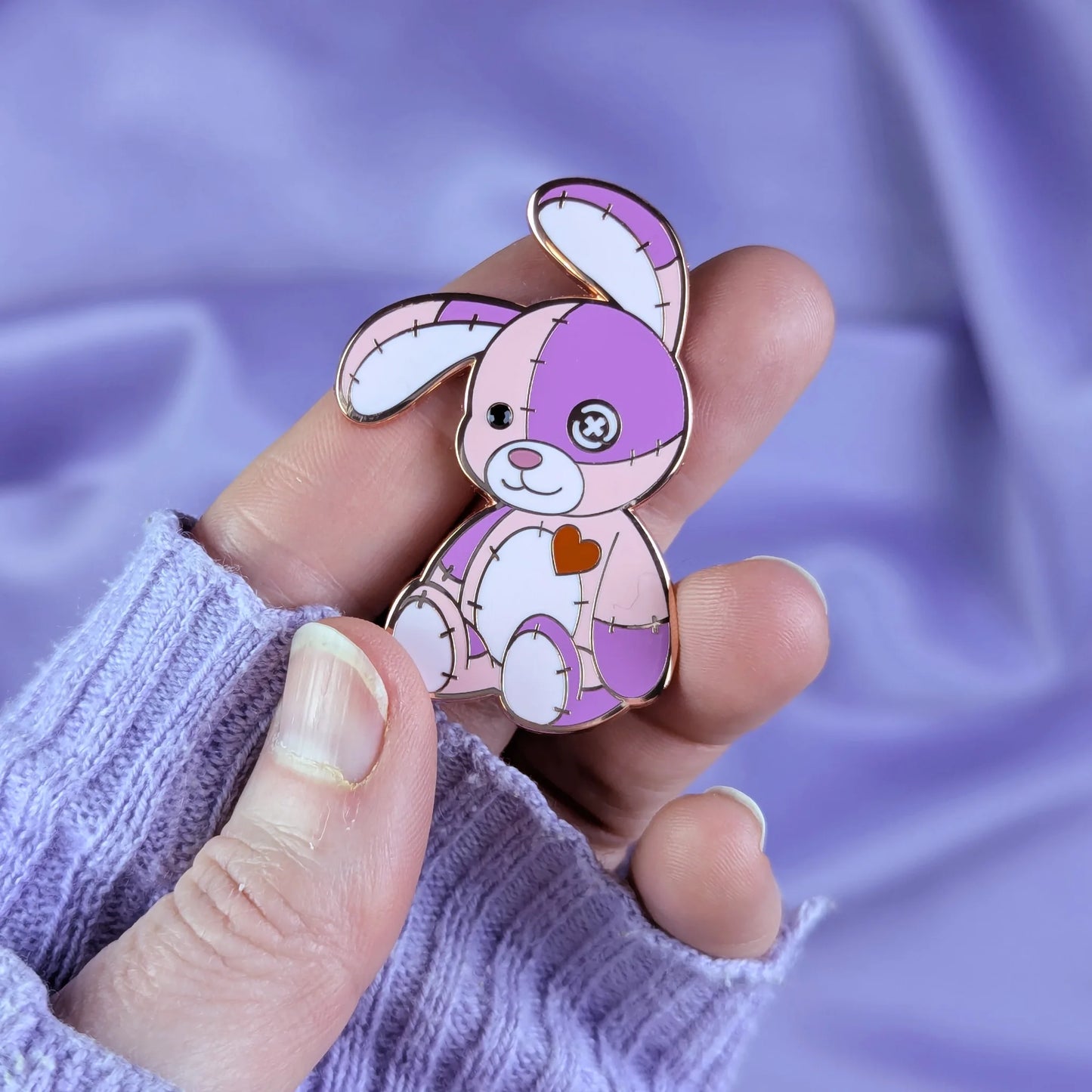 Hand holding a cute pink and purple patchwork bunny pin against a soft purple fabric background.