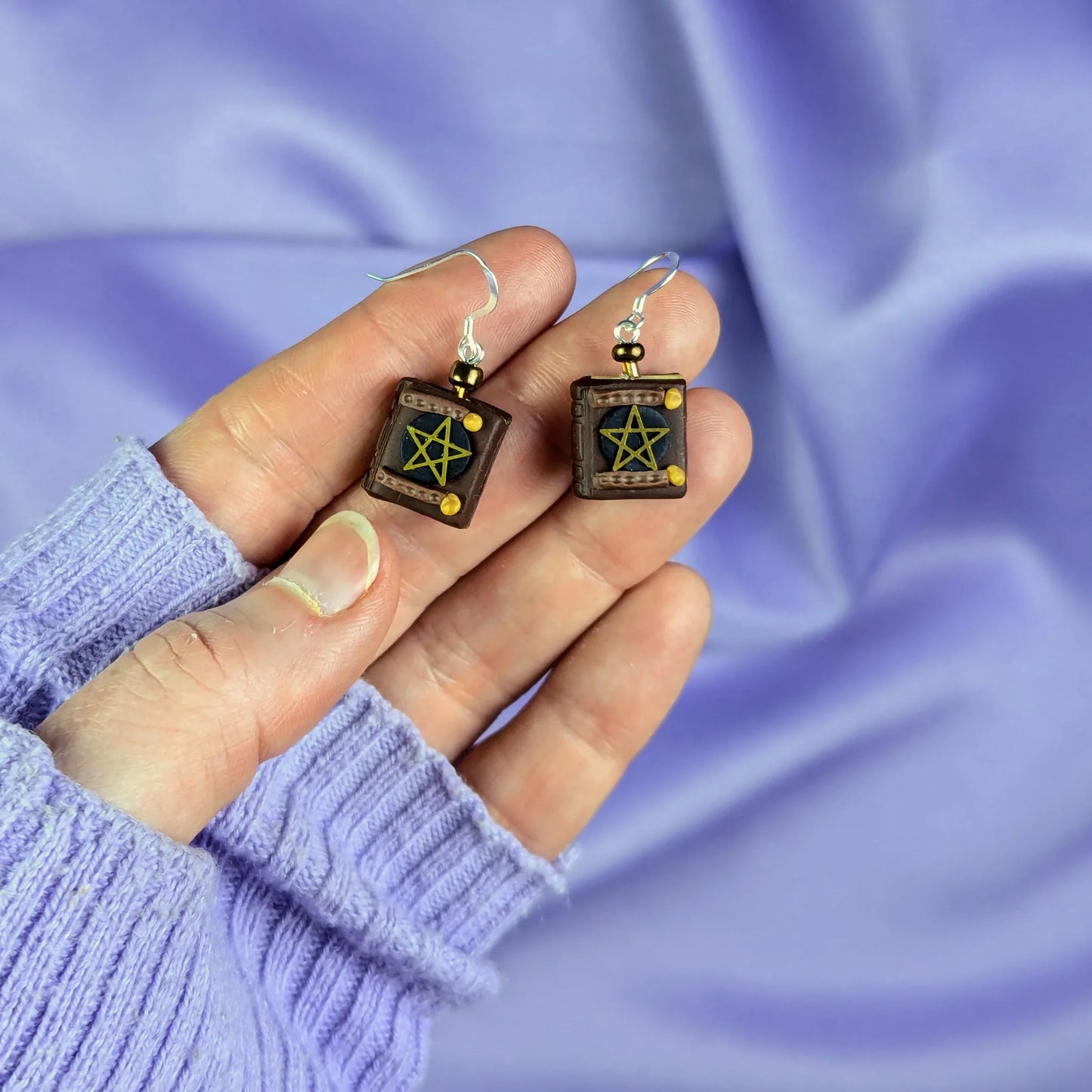 Hand holding two polymer clay earrings that resemble old ghost hunting books against a purple fabric background