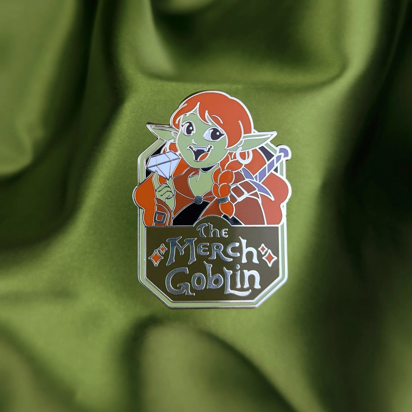 Hard enamel pin of a happy green goblin woman with long red hair, wearing armor and holding a diamond with text that says The Merch Goblin