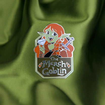 Hard enamel pin of a happy green goblin woman with long red hair, wearing armor and holding a diamond with text that says The Merch Goblin