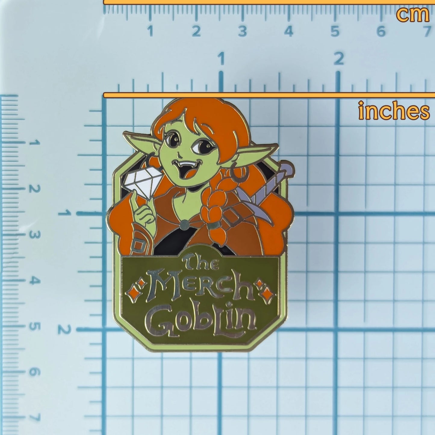 Pin with a goblin character and 'The Merch Goblin' text on a ruler grid background to show measurements.