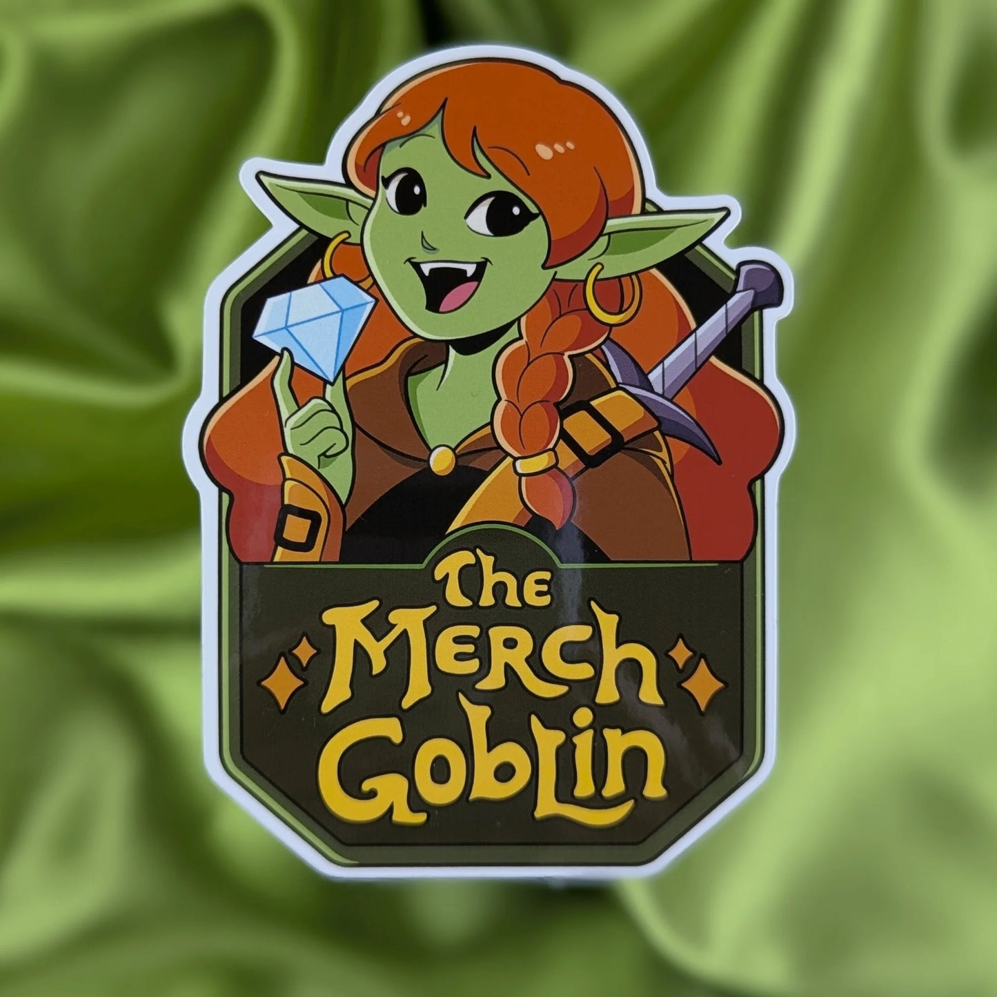 Sticker of a happy green goblin woman with long red hair, wearing armor and holding a diamond with text that says The Merch Goblin