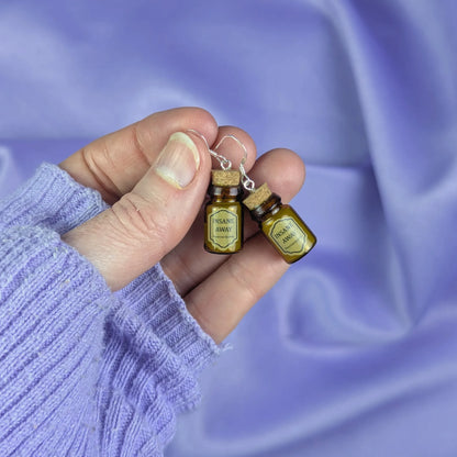 Hand holding a pair of miniature potion 
bottle earrings against a purple fabric background