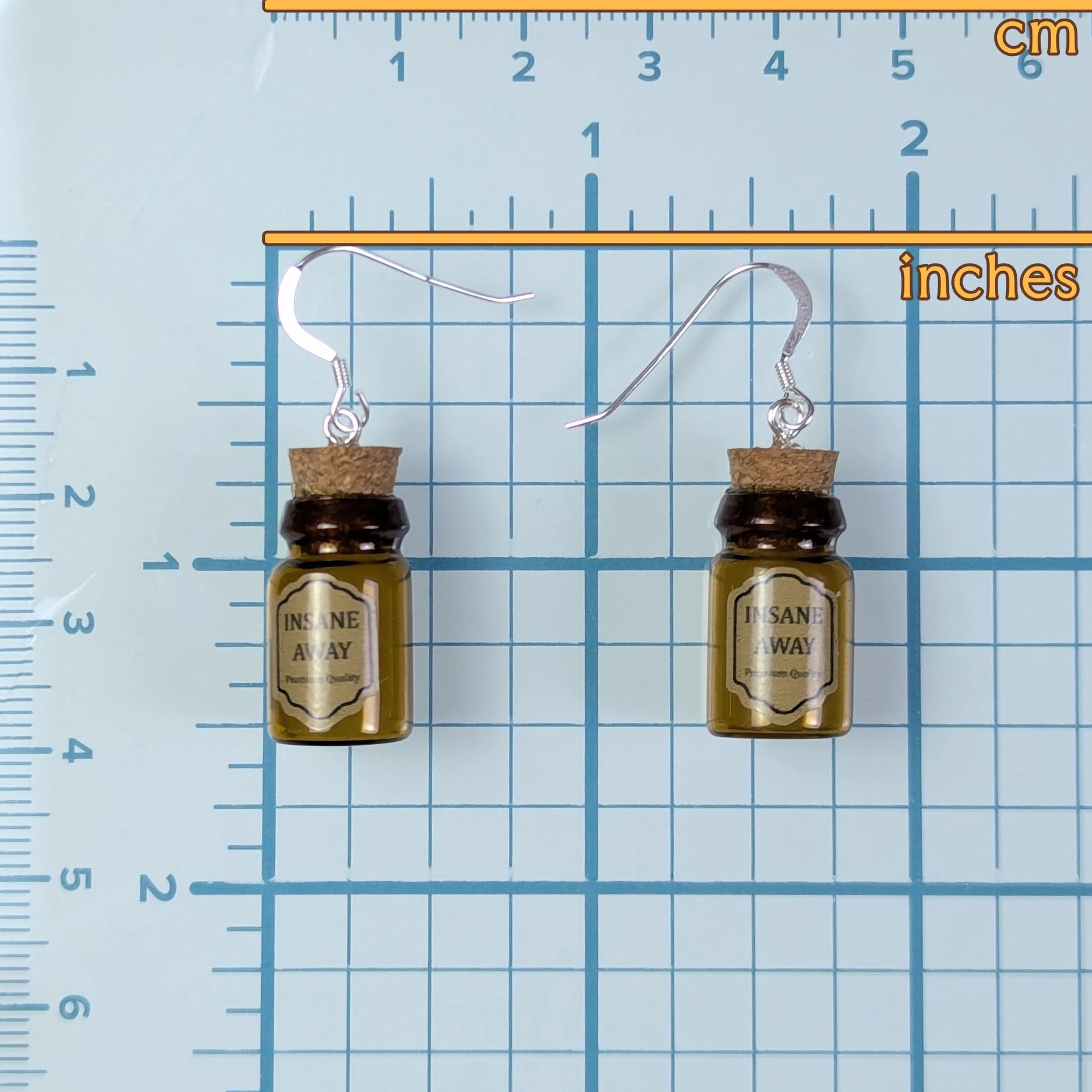 Pair of earrings resembling small bottles labeled 'Insane Away' on a blue grid background with measurements.