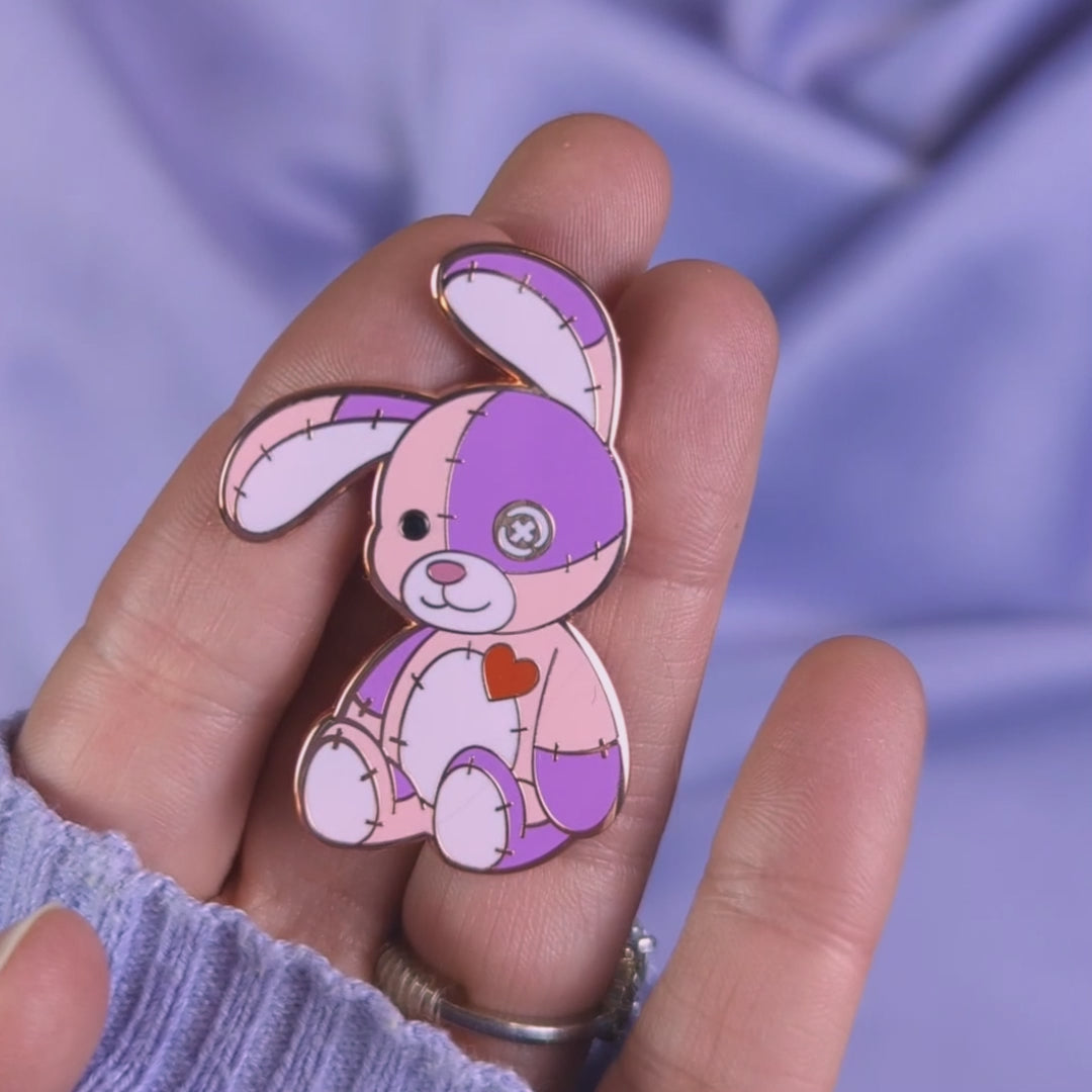 Video of pink and purple cute cursed stitched bunny hard enamel pin with rose gold metal, red heart, and button eye