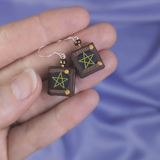 Video of a hand holding two polymer clay earrings that resemble old ghost hunting books against a purple fabric background