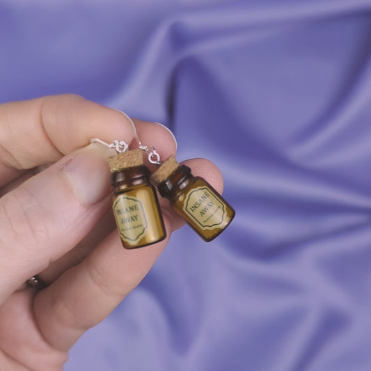 Video of a hand holding a pair of miniature potion bottle earrings against a purple fabric background