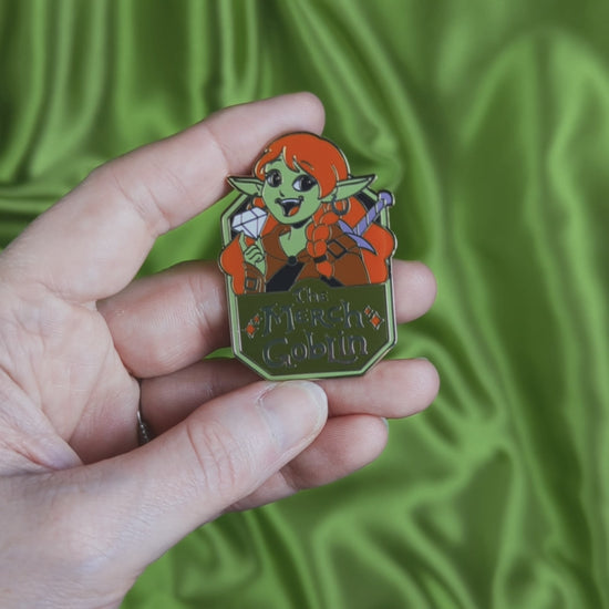 Video of a hard enamel pin of a happy green goblin woman with long red hair, wearing armor and holding a diamond with text that says The Merch Goblin