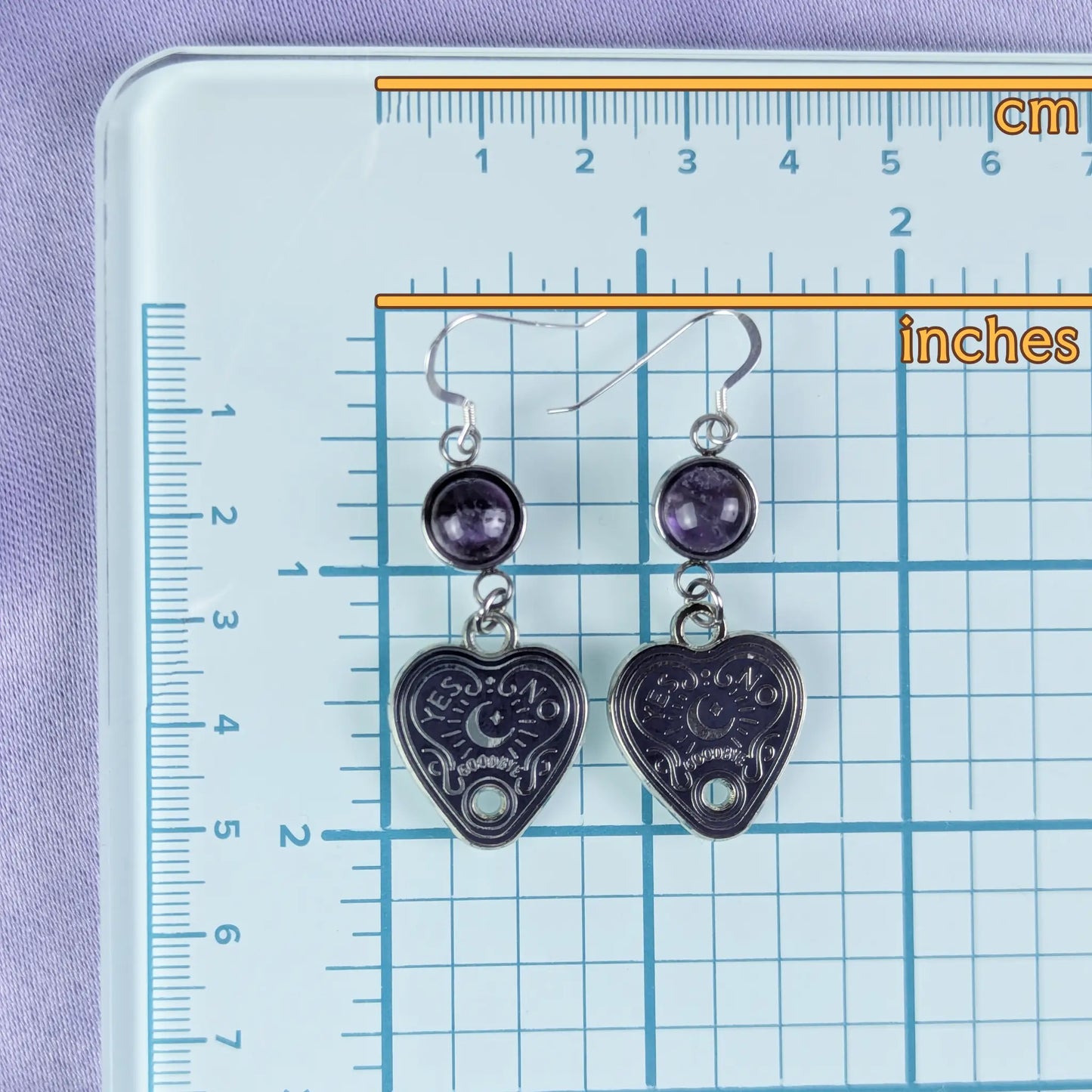 Heart-shaped earrings with purple stones on a ruler for scale against a purple background
