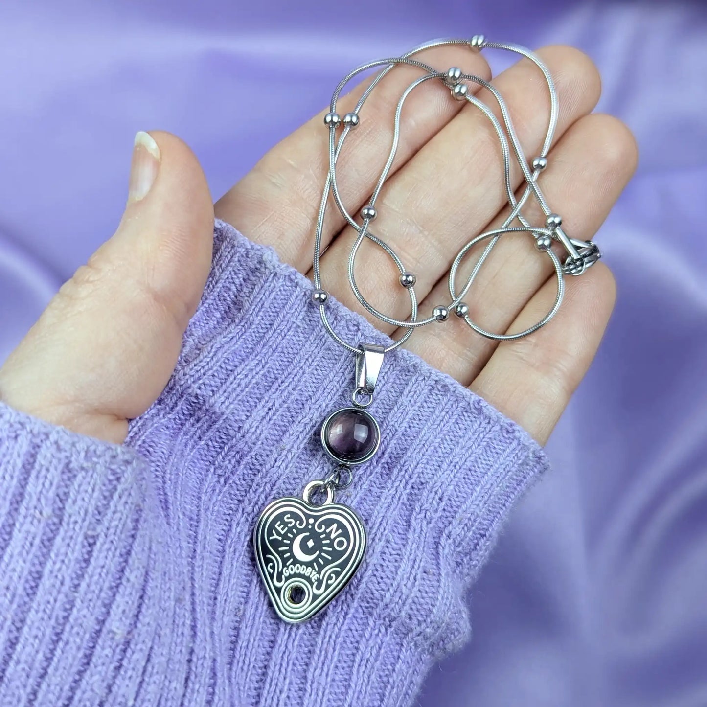 Stainless steel necklace with heart-shaped spirit board pendant and purple amethyst gemstone held in a hand