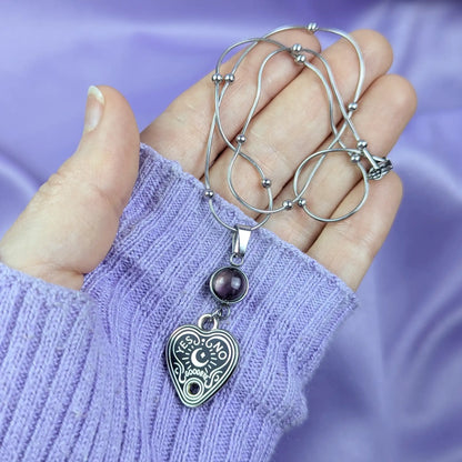 Stainless steel necklace with heart-shaped spirit board pendant and purple amethyst gemstone held in a hand