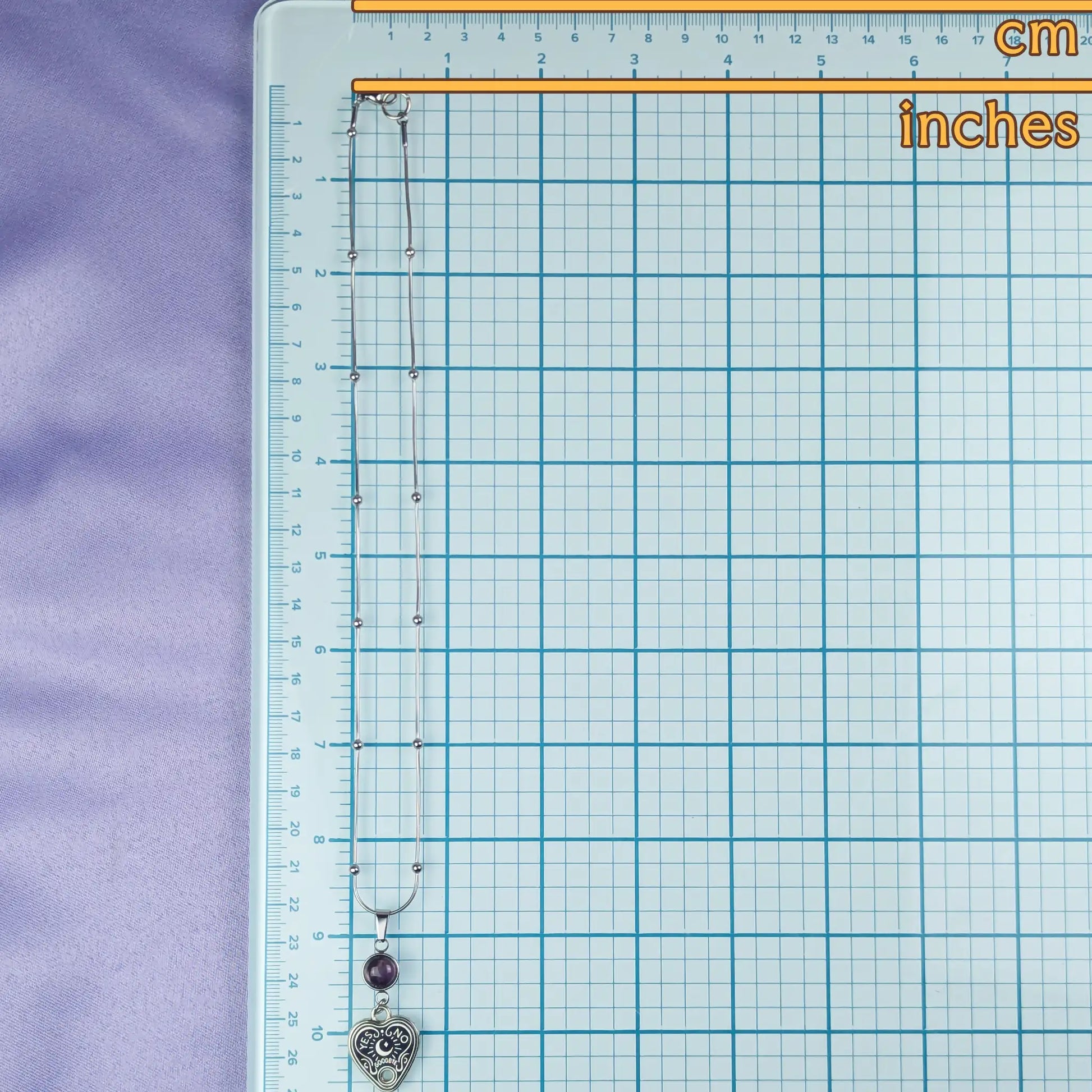 Necklace with heart-shaped pendant on a grid background with measurements in cm and inches.